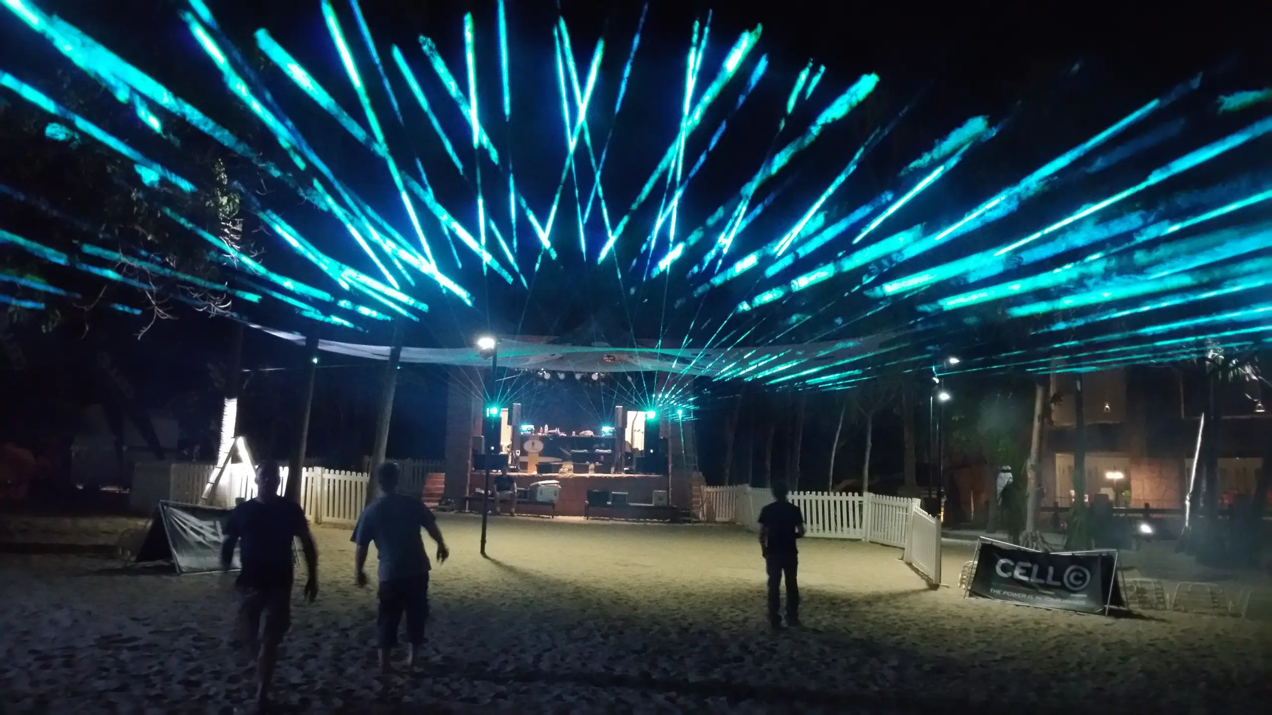 A fan of cyan laser beams over an outdoor stage at night, people walking towards it