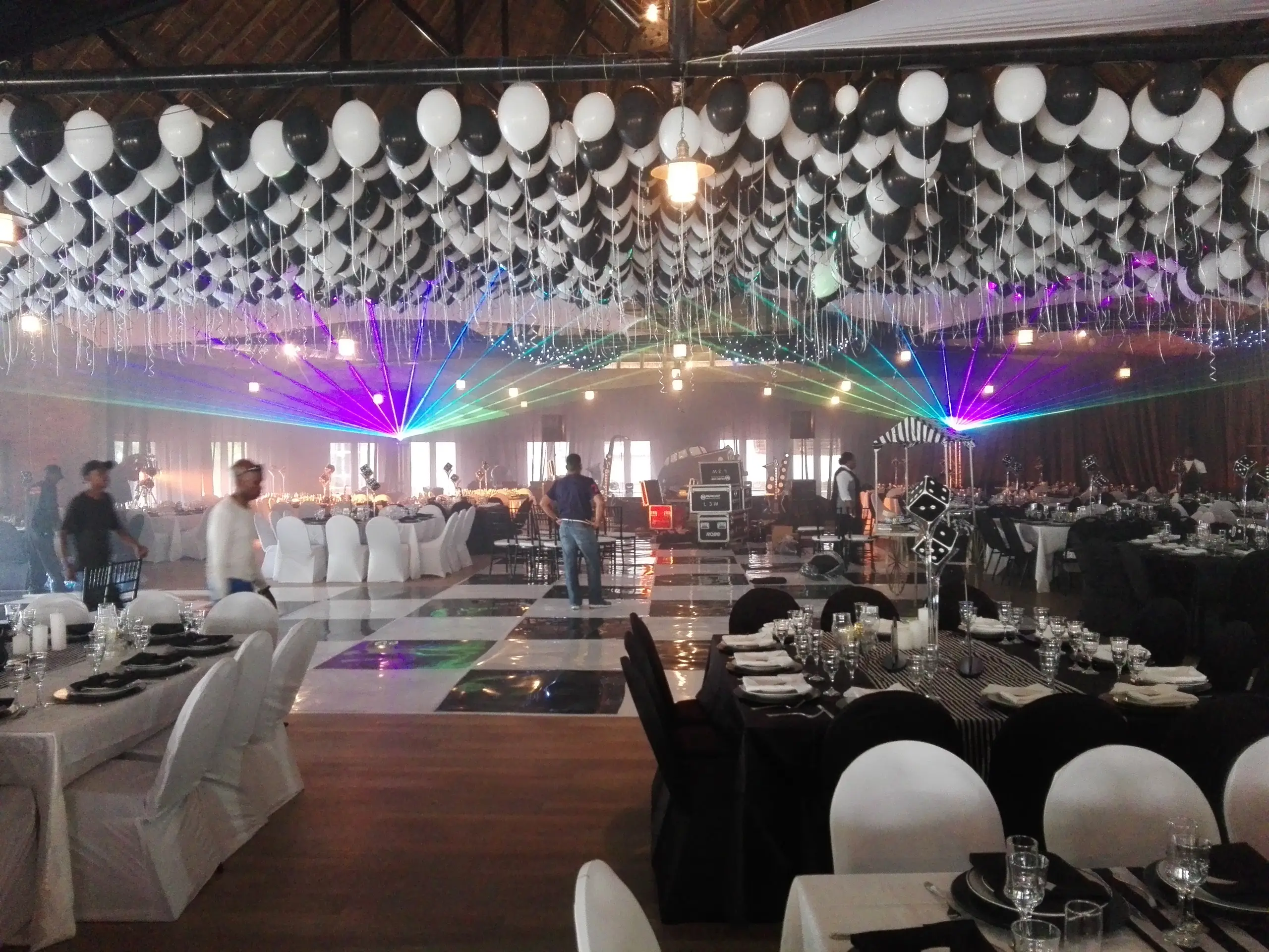 A function hall with a ceiling of white balloons, a black and white dance floor and lasers