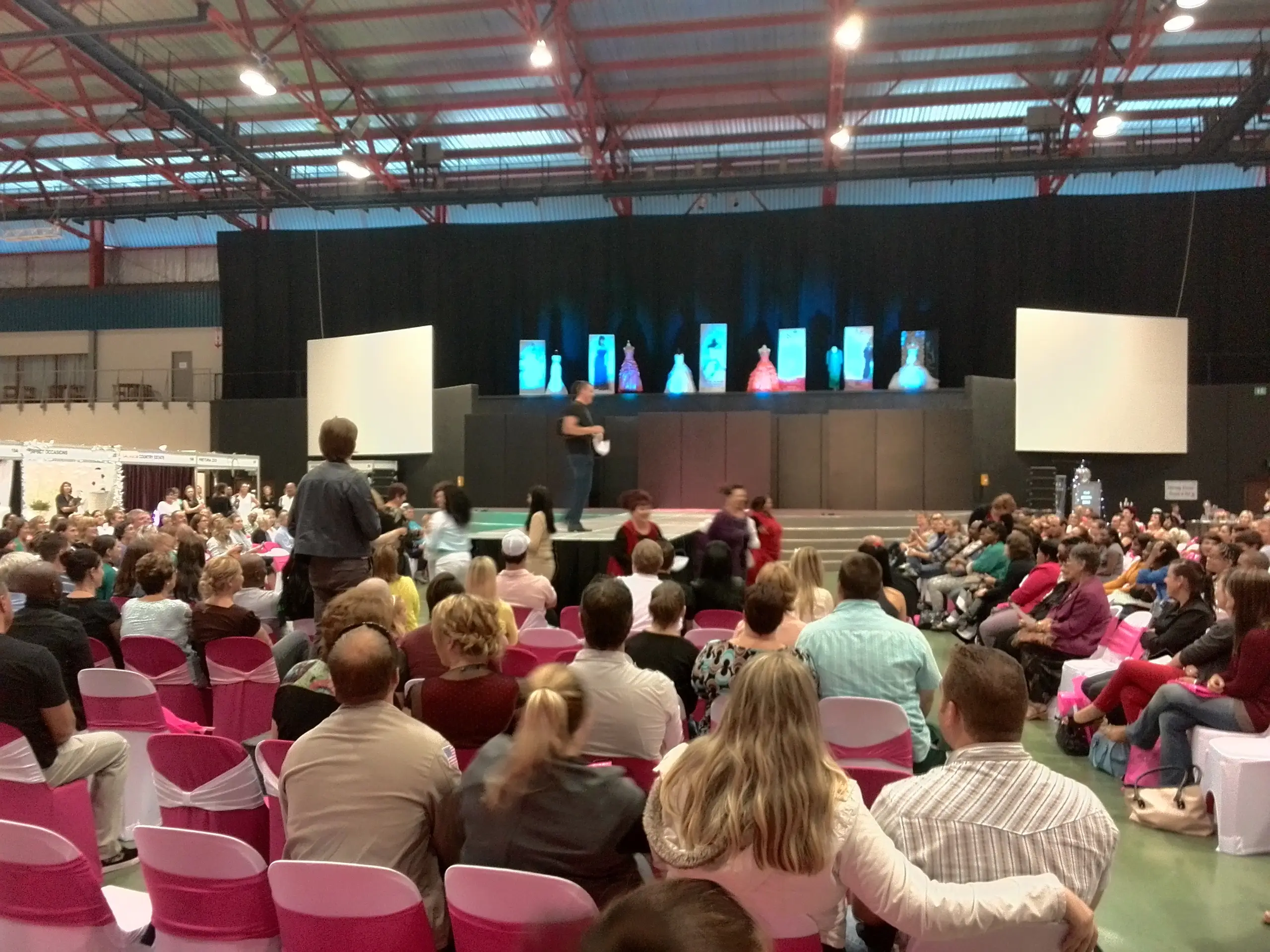 A fashion show runway in an exhibition hall with screens on either side and a seated audience