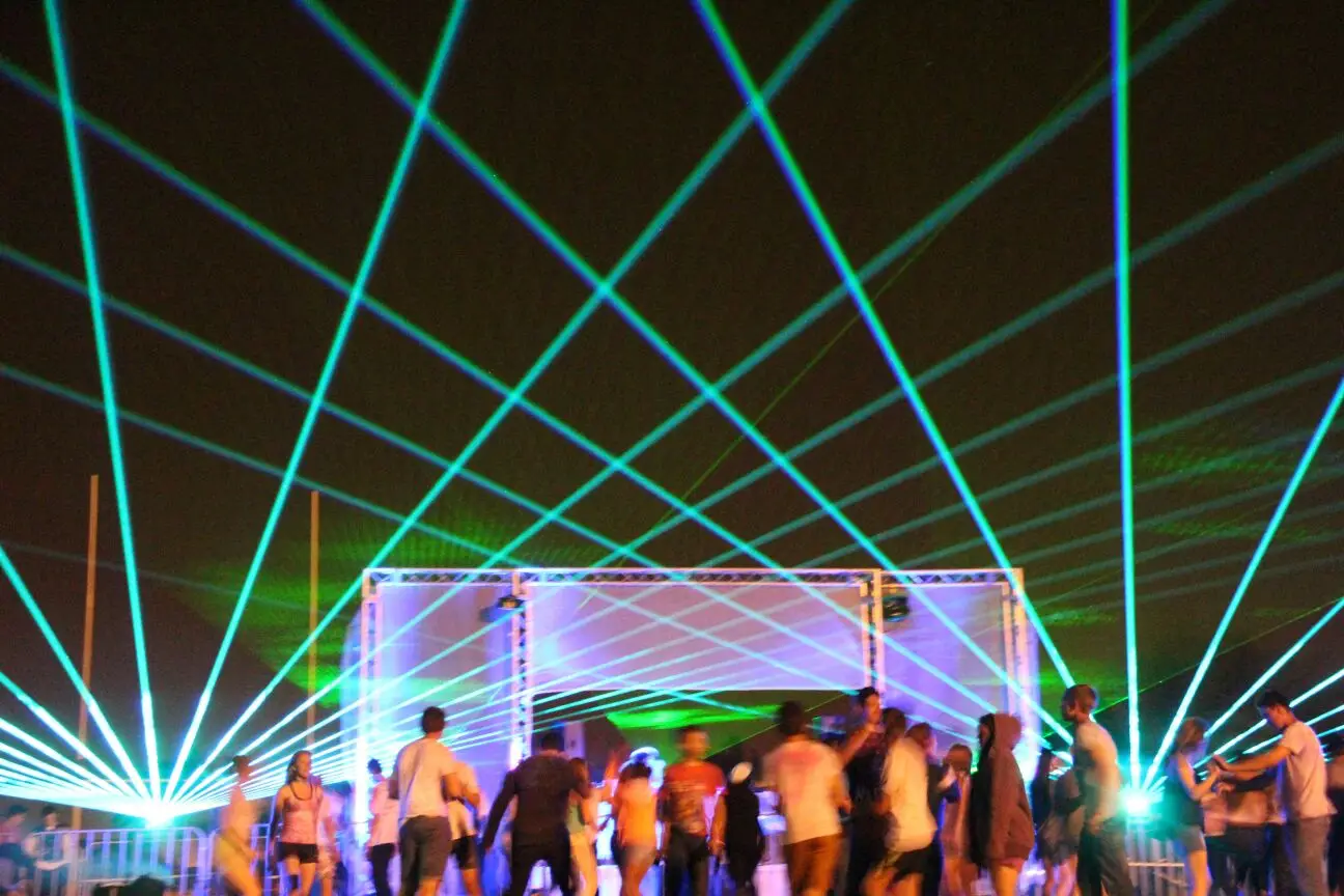 Green laser beams criss-crossing above a crowd dancing in front of a lit truss stage