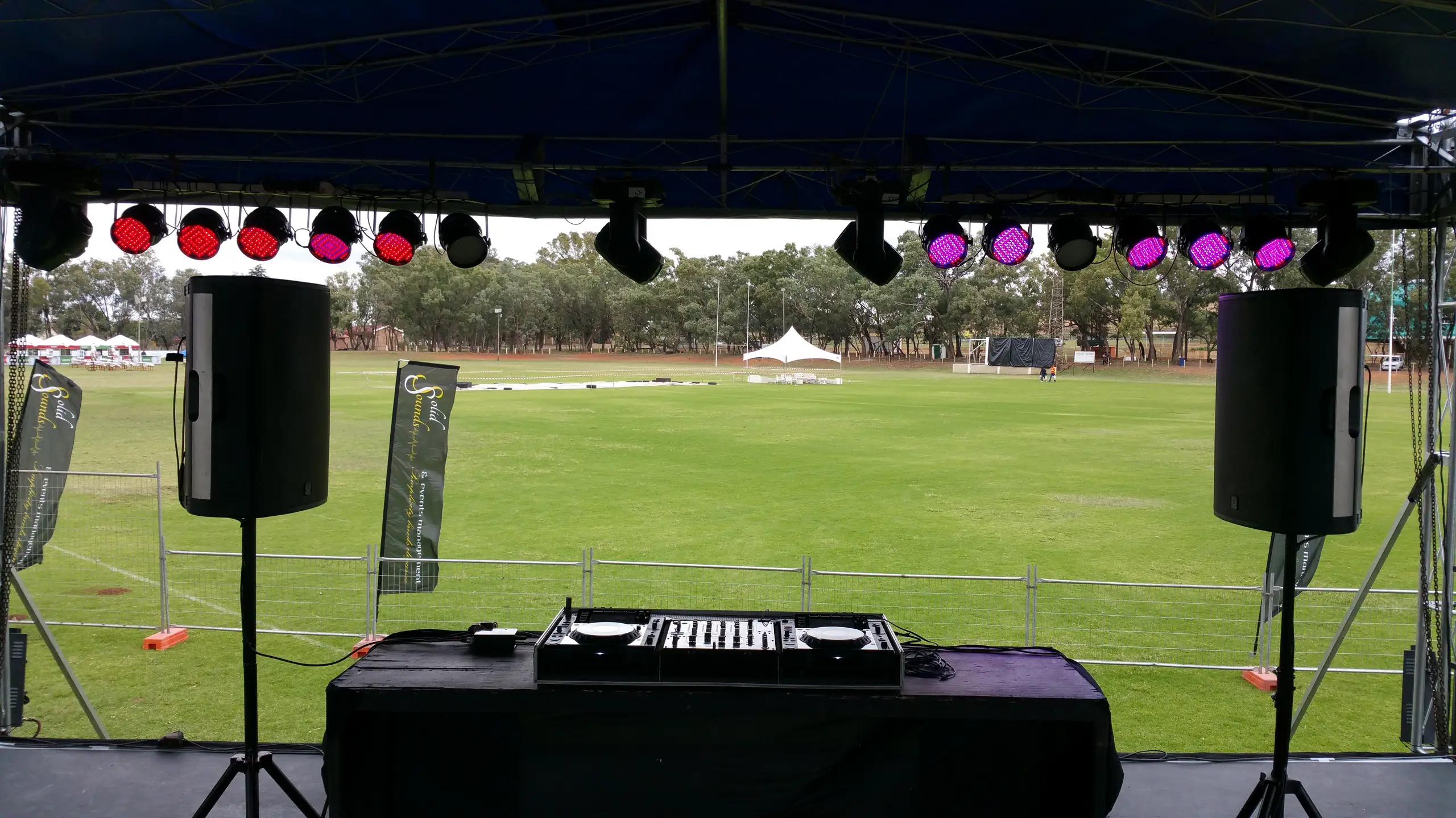 A stage roof with lights and speakers looking out over a sports field, DJ decks in front